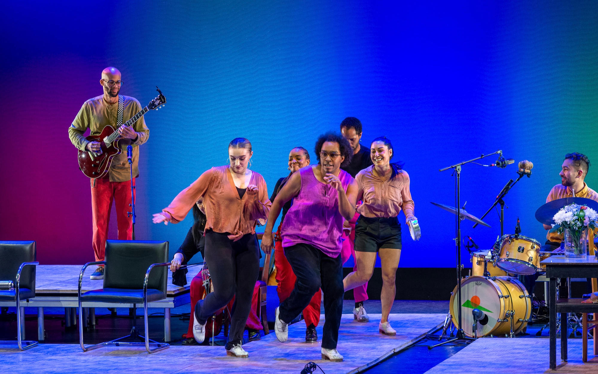 Dancers in bright shirts perform on a blue-green stage with musicians behind them