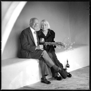 An elderly couple sitting closely together, holding a wine glass and a decorative branch.