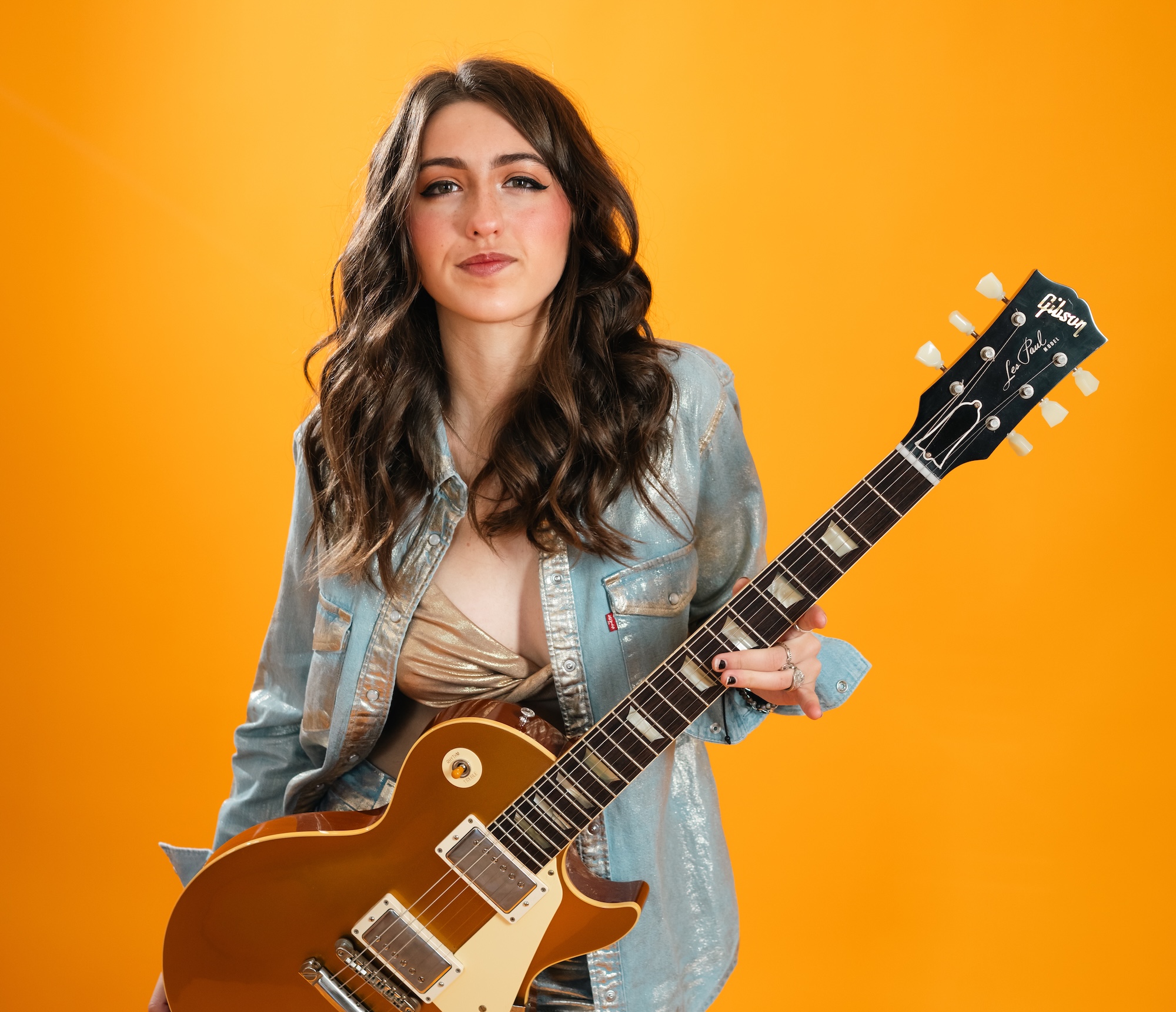 Bella Rayne holding brown electric guitar against bright orange background
