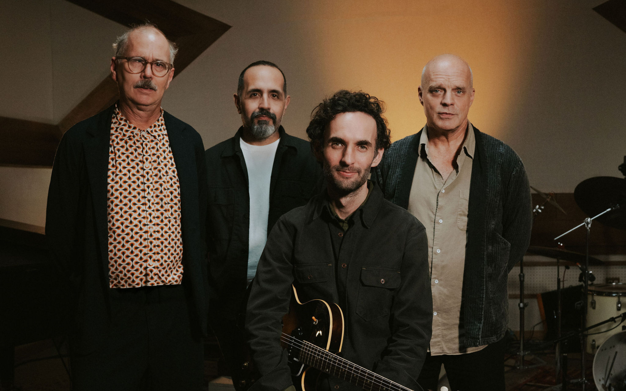 Julian Lage Quartet with John Medeski, Jorge Roeder, & Kenny Wollesen standing behind Julian Lage who is sitting and holding an electric guitar.