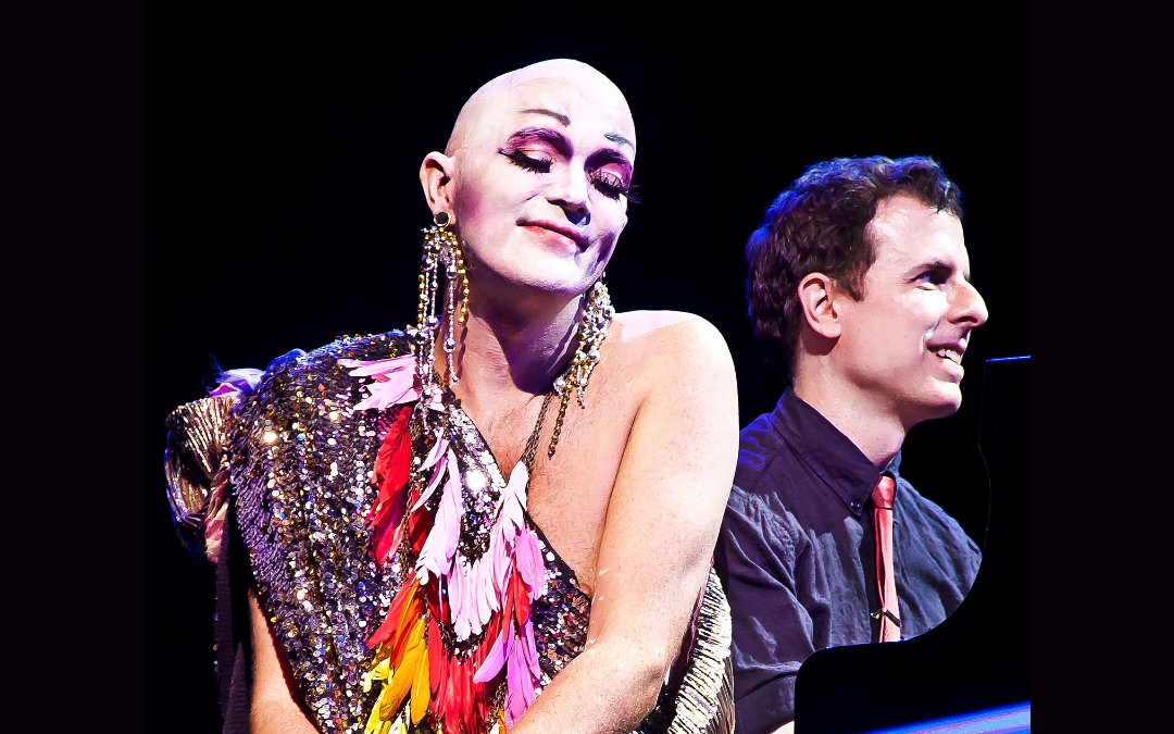 Taylor Mac wears a colorful costume smiles while pianist Matt Ray plays beside them.