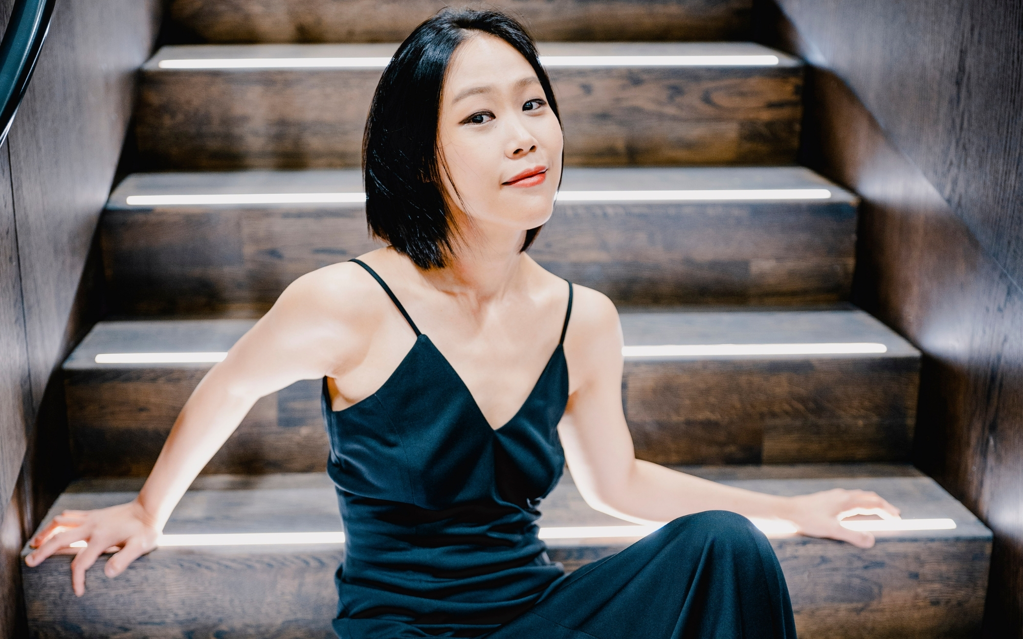 Woman in dark green dress seated on wooden stairs, relaxed posture, direct gaze.