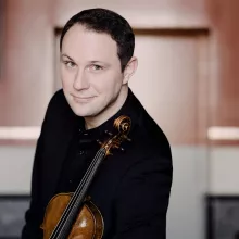 Smiling Jeffrey Myers in black holding a violin, warm indoor portrait
