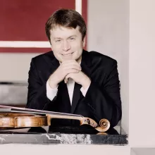 Jeremy Berry in black suit leans on ledge with violin
