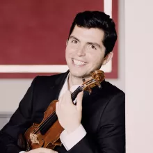 Ryan Meehan in suit holding a violin by a red wall (photograph)