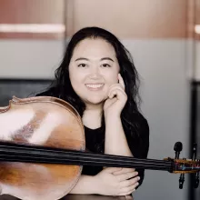 Estelle Choi resting chin on hand beside cello, warm studio background