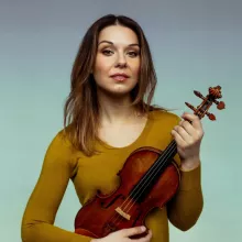 Amy Schroeder in a yellow sweater holding a violin, with a blue gradient background.