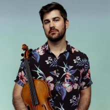Nathan Schram stands holding a violin, wearing a floral shirt against a blue gradient background.