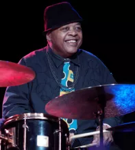 Jeff "Tain" Watts smiling, wearing a black hat and colorful tie, playing on stage.