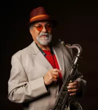 Joe Lovano wearing a fedora and sunglasses, holding a saxophone, against a dark background.