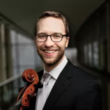 Nathan Vickery, smiling, in a suit holding a cello in a corridor