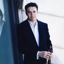 Noah Bendix-Bagley in dark suit holding a violin by reflective window (photograph)