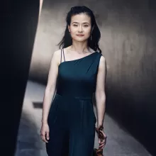 Shanshan Yao in a dark one-shoulder dress holding a violin in a narrow corridor