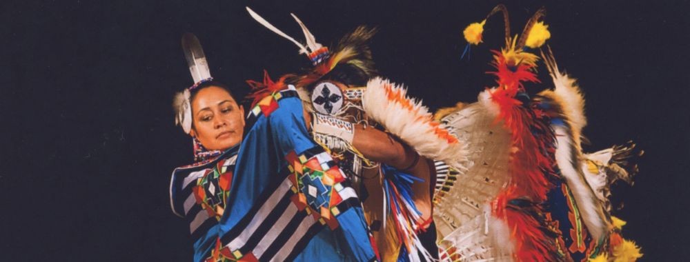 Two dancers in colorful feathered regalia performing against a dark background