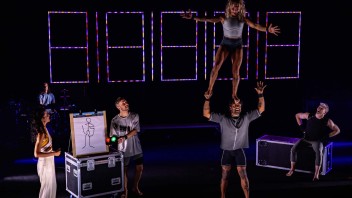 Acrobats performing on stage, one balanced on another’s shoulders, under colorful lights.