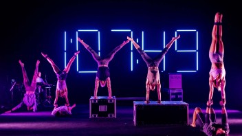 Acrobats performing handstands on stage with vibrant neon lights in the background.