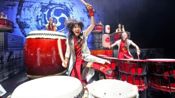 Two energetic taiko drummers in colorful costumes on stage with large red drums.