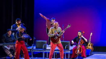 Five musicians on stage playing various instruments with a tap dancer behind them.