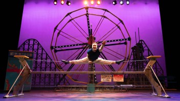 Acrobat performing a full split on a balance beam, purple-lit stage with Ferris wheel backdrop.