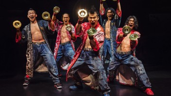 Six male Yamato performers in red jackets striking energetic pose holding gold cymbals on a dark stage