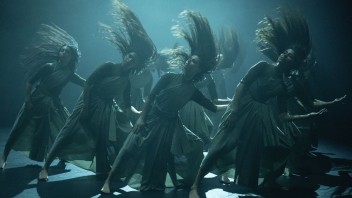 Multiple dancers in flowing gowns, hair and fabric mid-swish under moody blue-green stage lighting