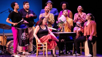 Diverse cast singing on stage, some playing percussion instruments, around a piano