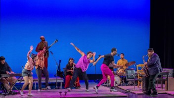 Musicians and dancers perform energetically on stage against a vivid blue backdrop