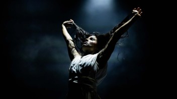 Woman onstage in dramatic spotlight, arms raised holding her hair.