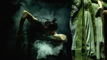 Dancers on a dark stage, one bowing amid green light and swirling smoke