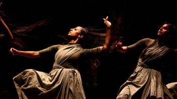 Female dancers in flowing gray dresses under dramatic warm spotlight