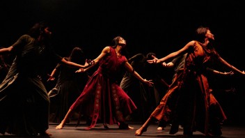 Dancers in flowing red-brown costumes on dimly lit stage, photograph capturing extended arms