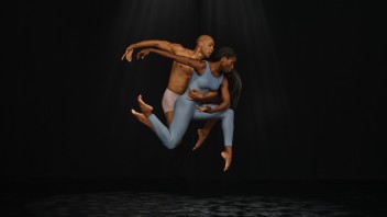 A man and a woman gracefully leap through a spotlight against a dark background.