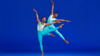 Two male dancers in turquoise leotards mid-leap against a deep-blue background.