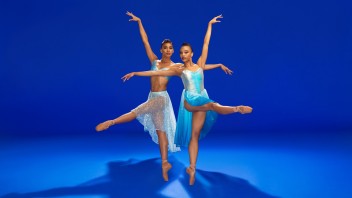 Two graceful ballerinas in blue and white costumes posing with their legs up away from each other against vivid blue backdrop