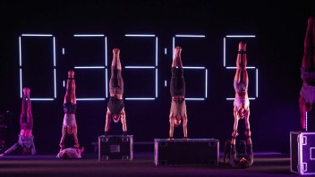 Acrobats perform handstands on stage, with a large digital timer in the background.
