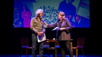 Two people on stage engage in conversation, with chairs and a colorful backdrop.