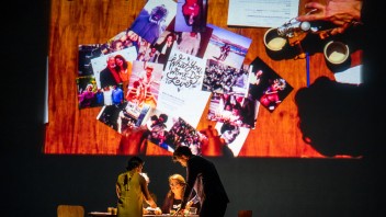 A wooden table topped with photographs and papers, with people discussing nearby.