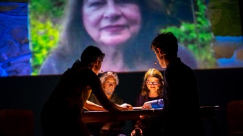 A group of people gathers around a table, with a woman’s face shown on a large screen behind them.