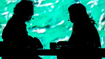 Two silhouetted figures sit at a table, facing each other, against a bright turquoise background.