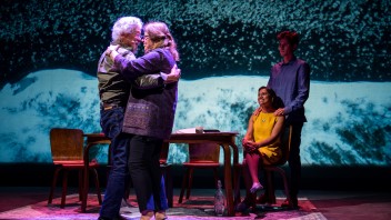 Two people embrace on stage, while two others sit at a table, with a colorful backdrop.