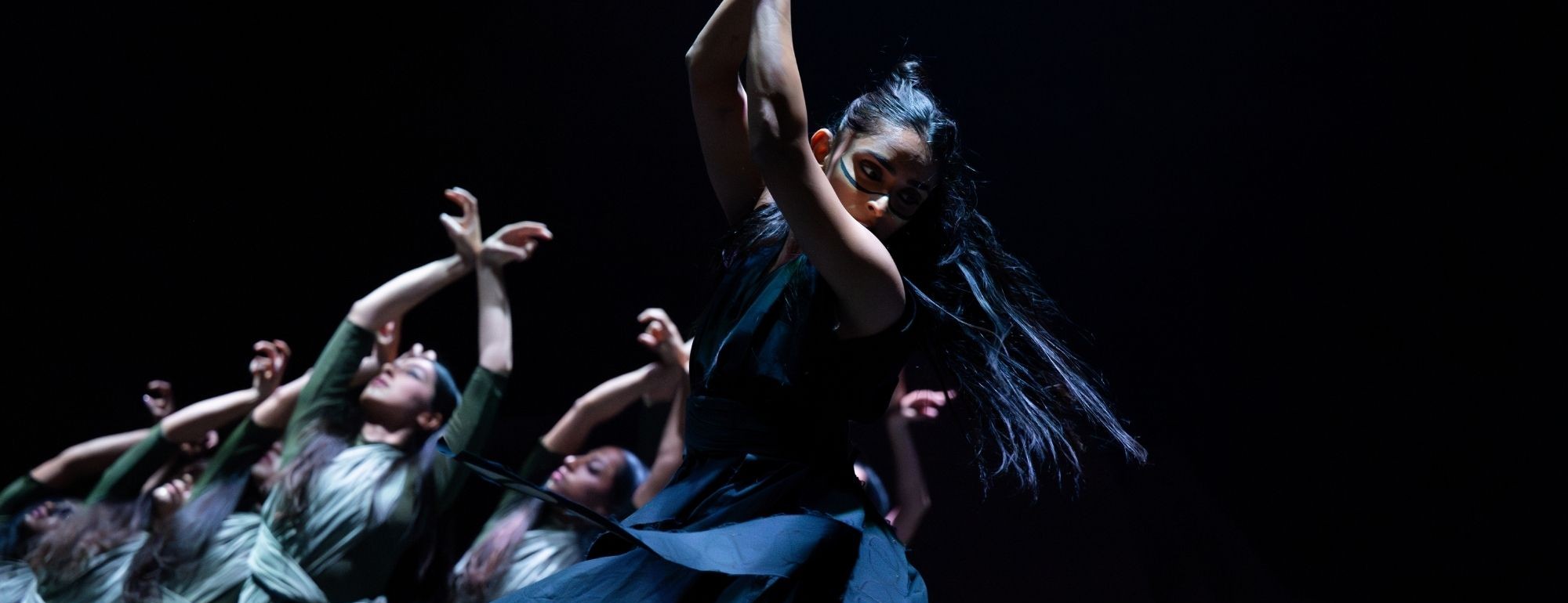 Dancers in teal dresses reaching upward on a dimly lit stage