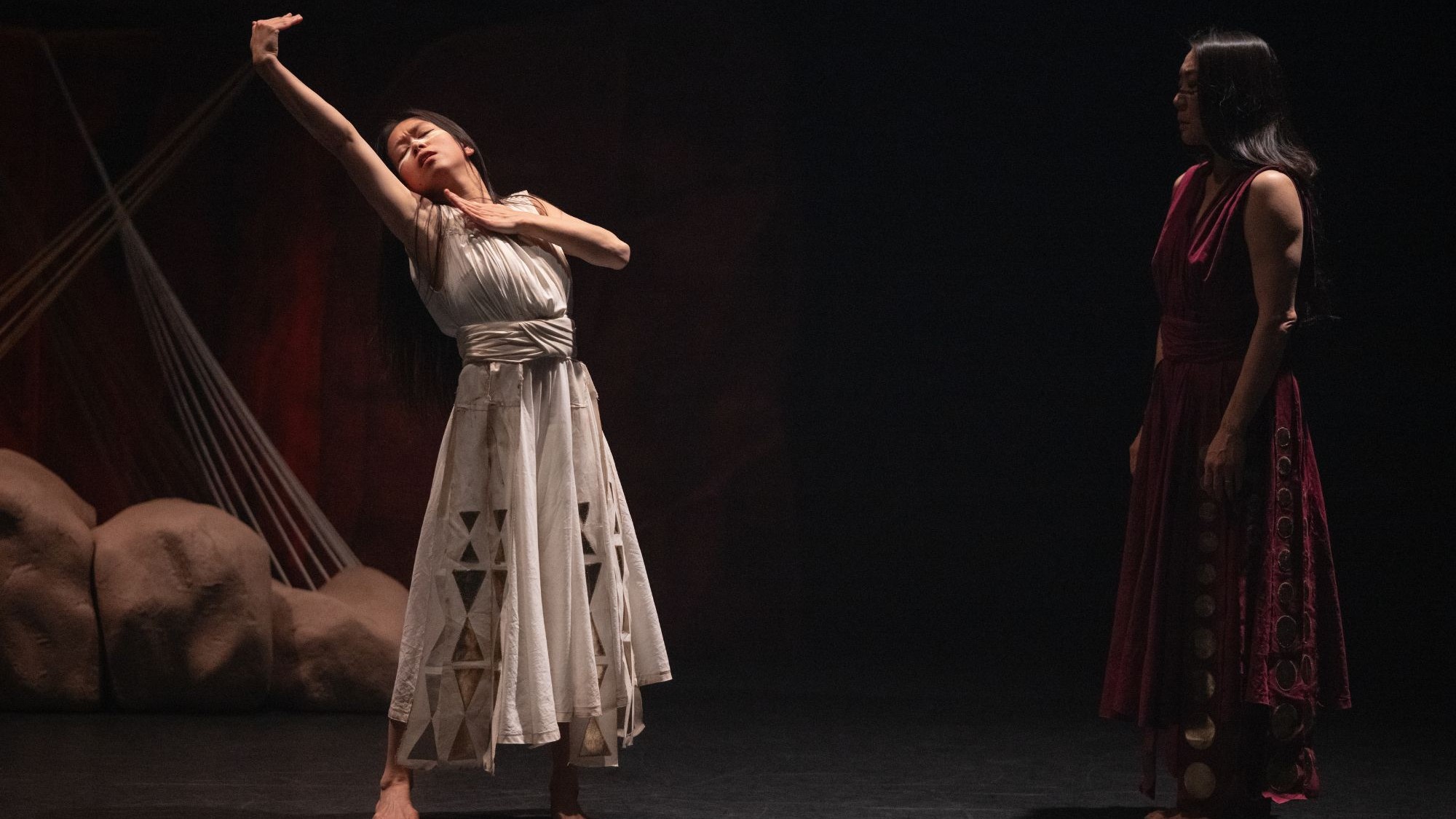 Two women on a dark stage; barefoot dancer in white dress reaching up, another watches.