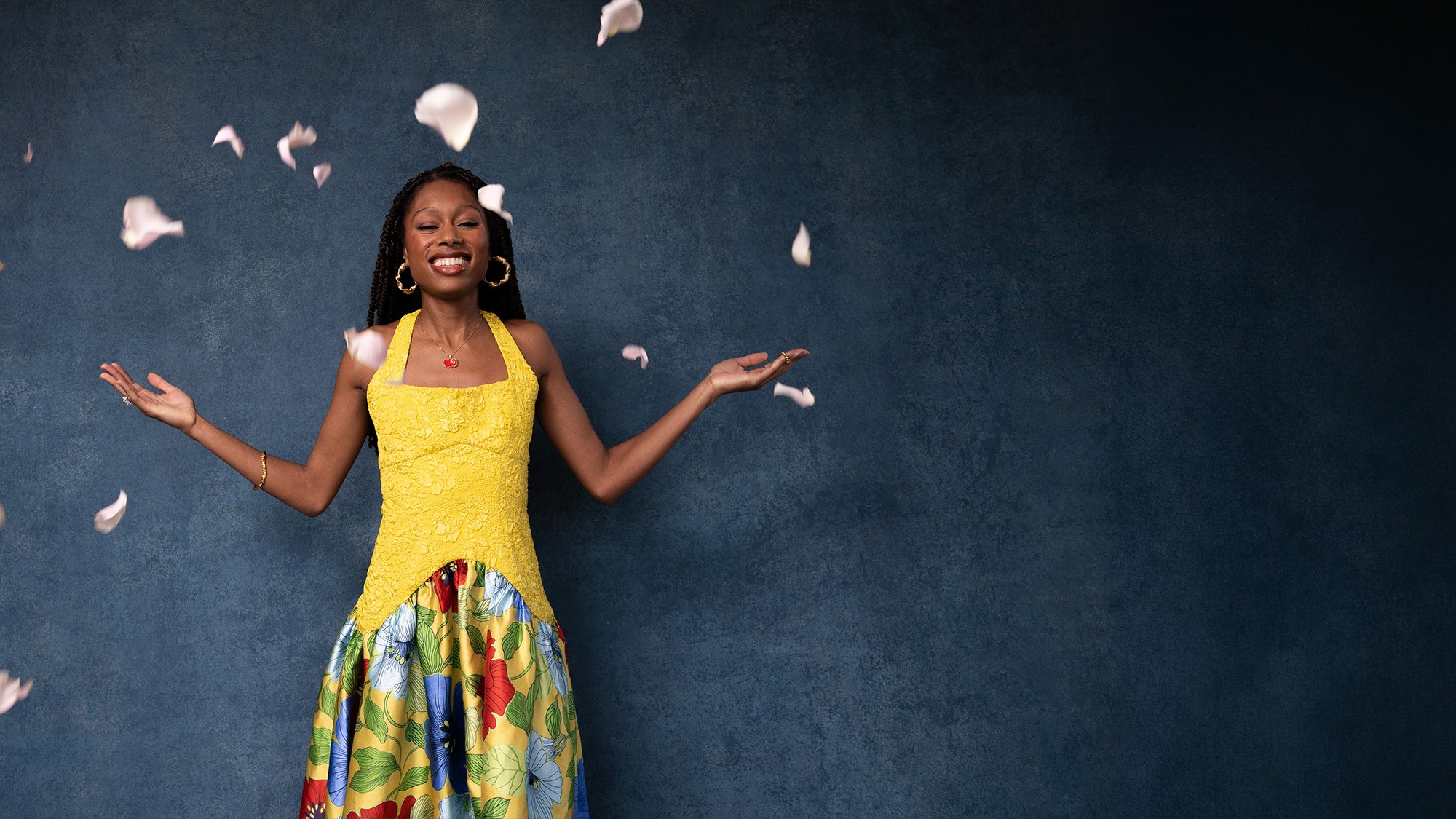 Isata Kanneh-Mason in yellow dress with patterned skirt tossing white petals against dark blue wall