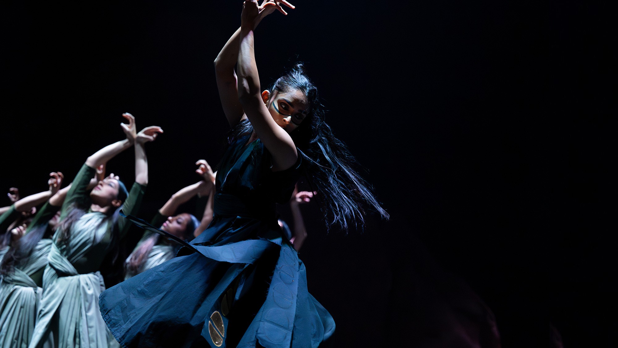 Group of dancers in flowing skirts reaching upward under moody blue stage lighting