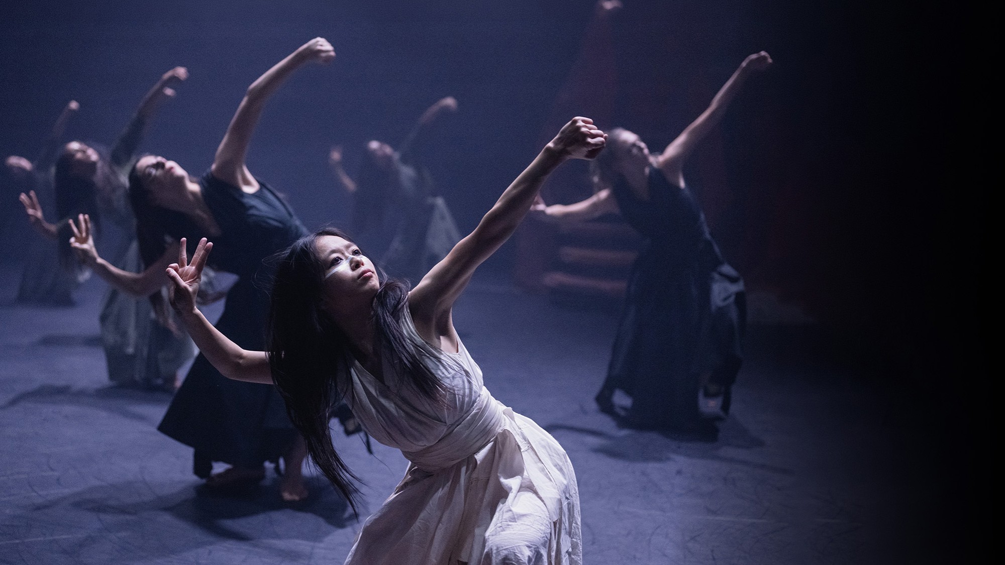 Lead dancer in white leans back amid troupe in dark dresses under moody stage lighting