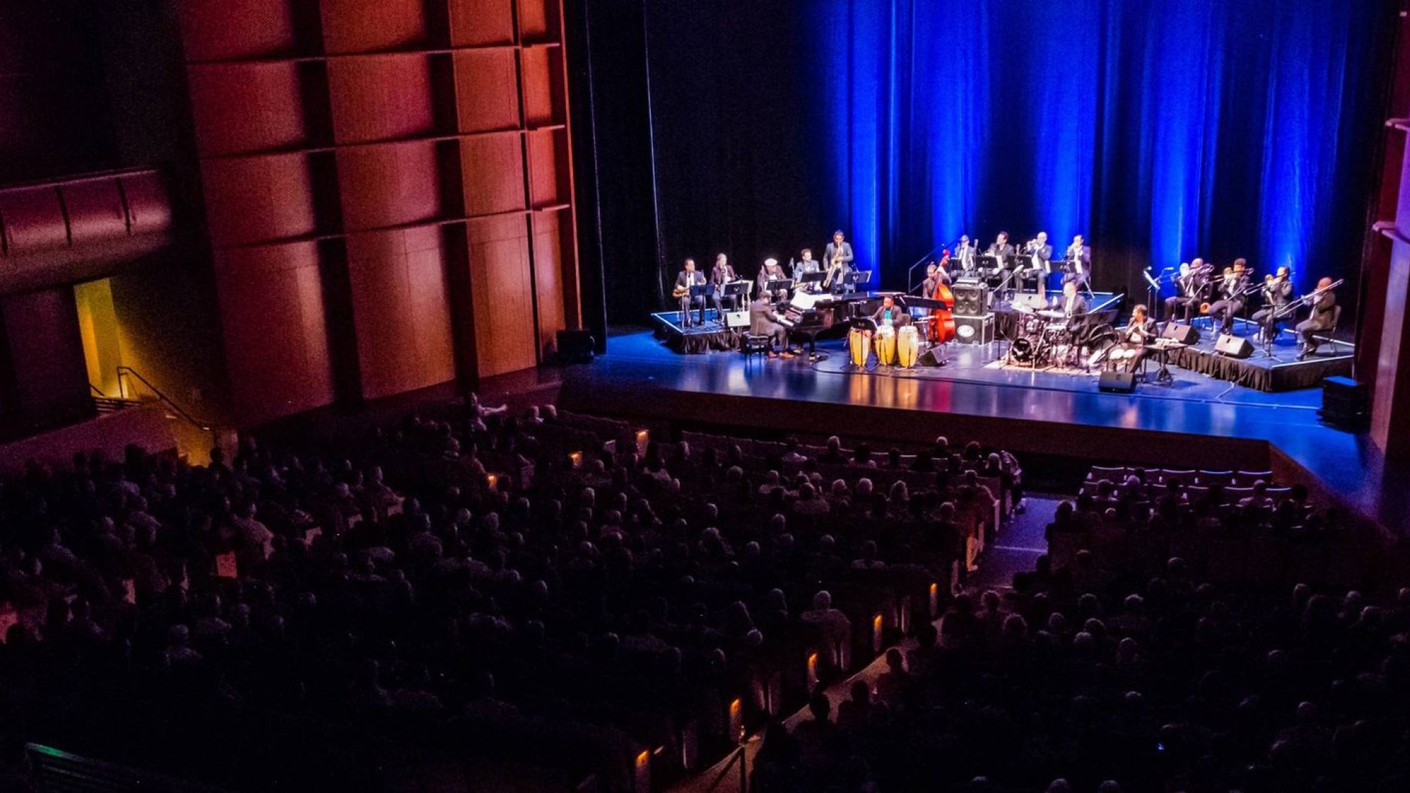 A large audience watches a musical performance on stage with colorful lighting.