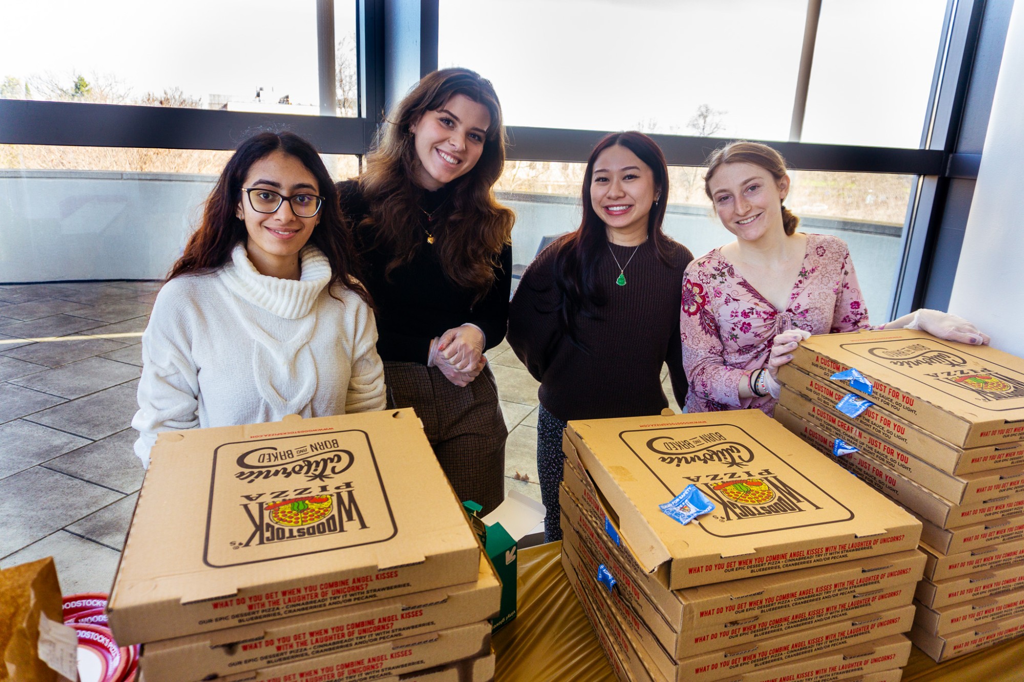 Four smiling students stand indoors near large windows, holding and stacking cardboard pizza boxes on a table. They face the camera, appearing to volunteer or distribute food, with additional pizza boxes and small condiment packets visible in front of them.