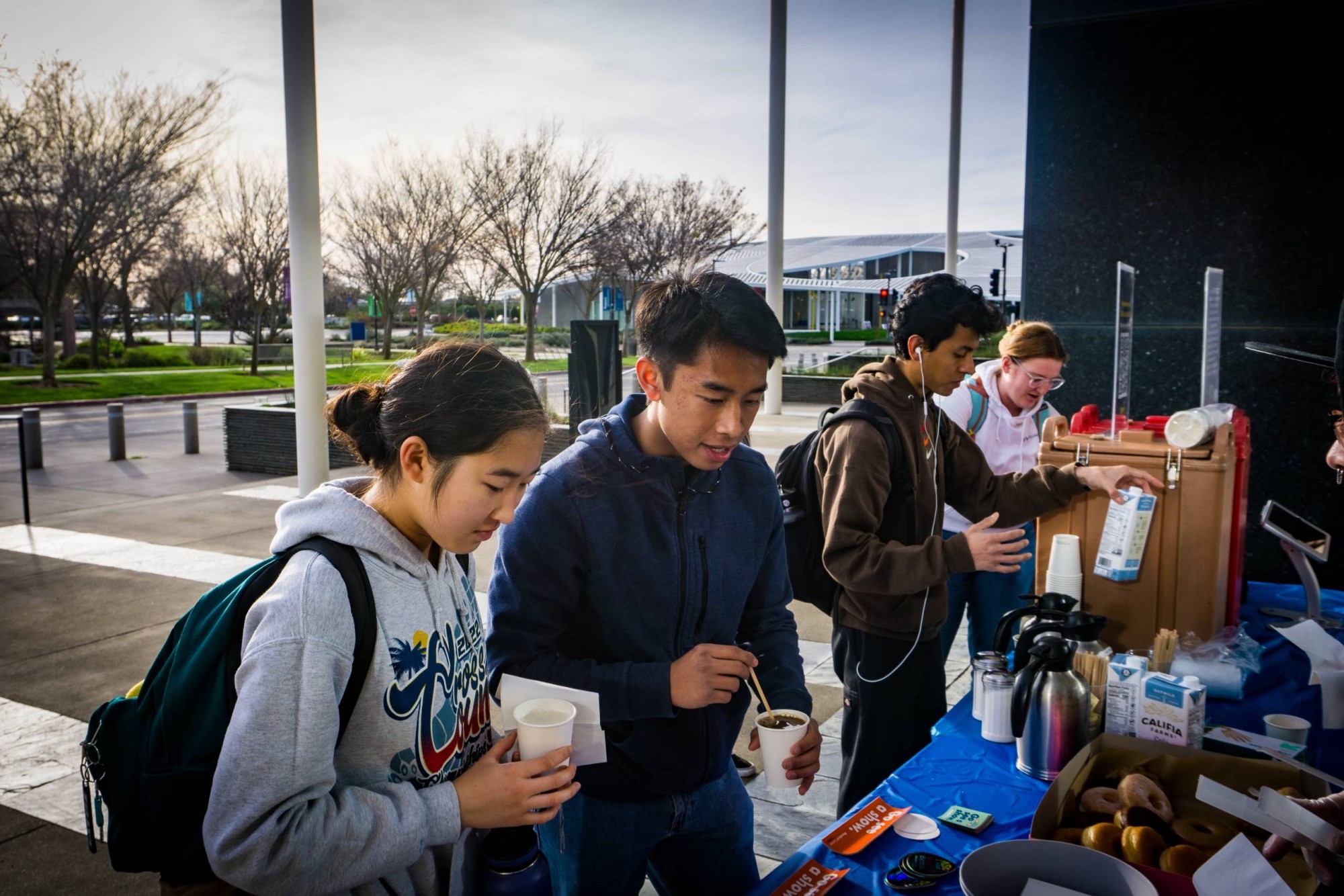 Several students stand outdoors at a refreshment table on a campus walkway, pouring and stirring hot drinks from insulated dispensers. They wear casual clothing and backpacks, with cups, creamers, and donuts arranged on a blue tablecloth, suggesting a free coffee or snack distribution event.