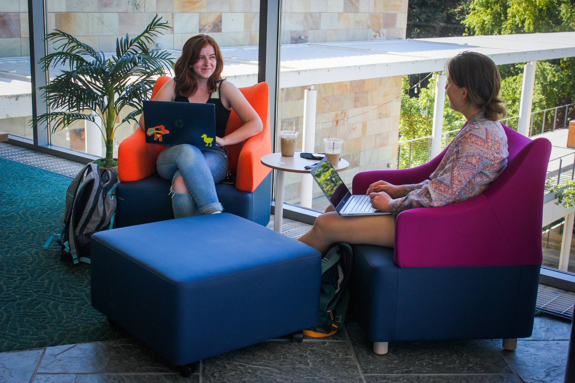 Two people sit facing each other in colorful lounge chairs inside a bright building with large windows. Each works on a laptop while chatting, with iced coffee cups on a small table between them and backpacks nearby, suggesting a relaxed study or work meeting in a campus common area.