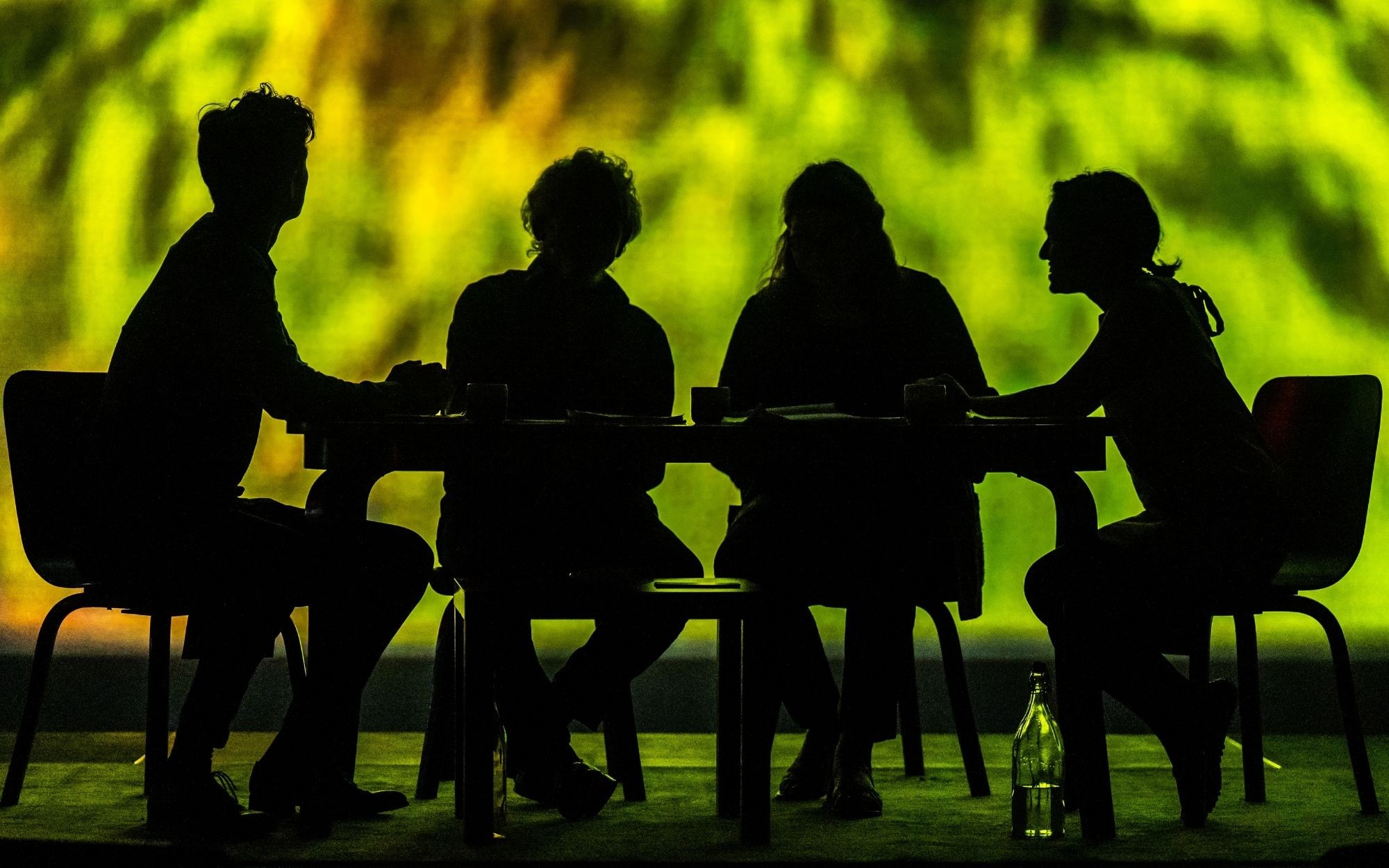 Four silhouetted people seated around a table against a vivid green-yellow backdrop